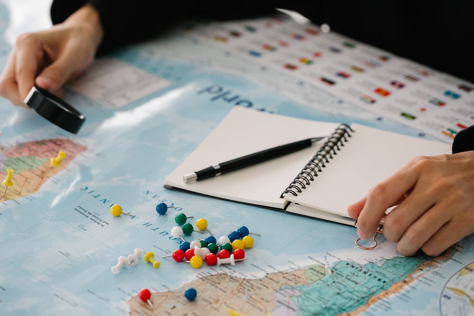 Hands placing pins on a world map beside a notebook, sketching a cruise itinerary template